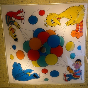 Pode incluir: Luminária quadrada com personagens da Vila Sésamo: Bert, Big Bird, Cookie Monster e Ernie. Os personagens seguram balões coloridos. A luminária tem um cabo branco.