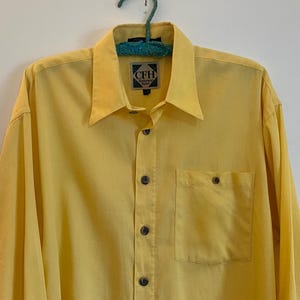 May include: A bright yellow button-down shirt with long sleeves, a chest pocket, and a collar. The shirt has dark buttons and a small label near the collar. The shirt is hanging on a green hanger.