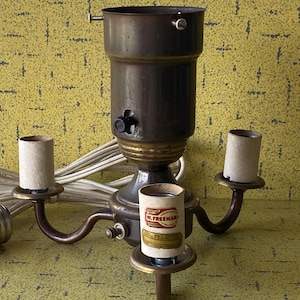 May include: A vintage brass and bronze lamp fixture with three arms for bulbs. The fixture has a central socket, a power cord, and three cardboard sleeves for the bulbs. The fixture is on a yellow background.