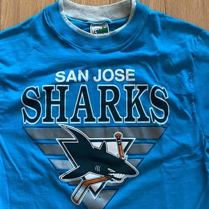 May include: A turquoise San Jose Sharks t-shirt with a grey collar. The shirt features the team name in white and a logo of a shark with a hockey stick and a triangle design.