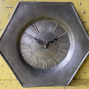 Peut inclure: Horloge murale hexagonale de couleur argentée avec des chiffres romains. Le cadran de l'horloge est circulaire, avec des aiguilles noires et un petit centre doré. Le cadre de l'horloge est argenté mat.