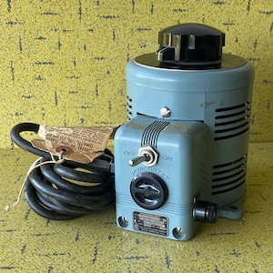May include: A vintage blue and black electrical transformer with a black knob on top and a switch labelled "ON" and "OFF". A black power cord is coiled next to the transformer, with a tag attached. The transformer has a dial labelled "POWERSTAT".