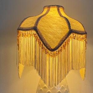 May include: A bell-shaped lampshade with a textured cream fabric surface, featuring white lace trim along the edges and a decorative fringe of long cream-coloured tassels interspersed with amber-coloured glass beads hanging from the bottom.