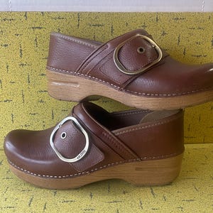 Può includere: Un paio di zoccoli in pelle marrone con suole in legno. Ogni zoccolo presenta una grande fibbia decorativa in tonalità oro o argento. Gli zoccoli hanno una punta arrotondata e un leggero tacco, adatti all'uso quotidiano.