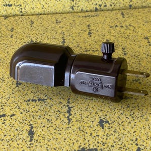 May include: A brown electrical plug adapter with a rotating knob and two prongs. The adapter has a rectangular shape with rounded edges and the text "7W 125V USA".