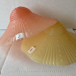 May include: Two vintage glass lampshades. One is a soft peach colour, the other is a pale yellow. Both have a scalloped edge and a series of small, evenly spaced circles. The peach shade has the number "2" on a white sticker, and the yellow shade has the number "1".