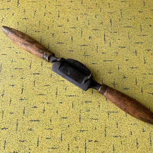 以下が含まれることがあります： 暗い金属の刃と2つの木製ハンドルを備えたヴィンテージの引きナイフ。ハンドルは明るい茶色で、金属リングで刃に取り付けられています。このツールは木材を成形するために設計されており、素朴な外観をしています。