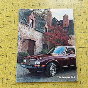 Op de afbeelding: Een vintage brochure met een donkerrode Peugeot 504 sedan geparkeerd voor een bakstenen huis met een steil dak en een boom met rode bladeren. De tekst The Peugeot 504 staat in het wit gedrukt in de rechterbenedenhoek van de papieren omslag.