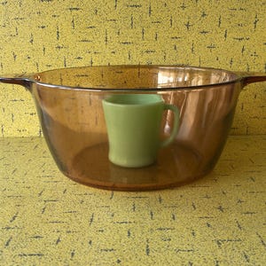 May include: An amber glass casserole dish with handles, containing a small, light green mug. The background is a yellow surface with a repeating pattern. The dish is suitable for cooking.