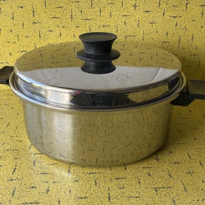 Puede incluir: Una olla de acero inoxidable con tapa y asas negras. La olla tiene una superficie brillante y reflectante y un pomo negro en la tapa. La olla está sobre una superficie amarilla.