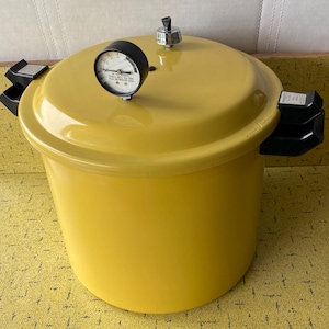 May include: A bright yellow pressure canner with a matching lid, featuring black plastic side handles, a circular pressure gauge on top, and a central metal vent pipe. The gauge face includes text indicating pressure settings for safe operation.