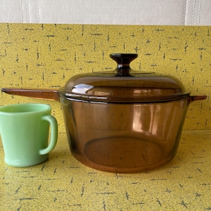 Può includere: Una pirofila vintage in vetro ambrato con coperchio e una tazza verde. La pirofila ha un manico ed è accanto a una piccola tazza verde. Gli oggetti sono su una superficie gialla.
