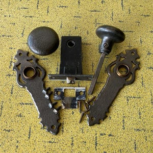 May include: A set of dark metal door hardware including two ornate rectangular backplates with circular openings, a round knob with a spindle, a rectangular latch mechanism, a strike plate, and several small metal screws for installation.