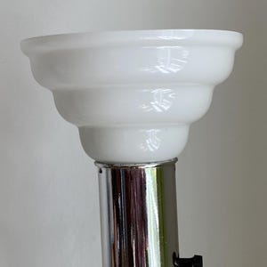 Op de afbeelding: Een witte glazen lampenkap met een gelaagd ontwerp staat op een chromen metalen paal. De kap heeft een glanzende afwerking en een afgeronde bovenkant. De lamp is waarschijnlijk voor de inrichting van het huis.