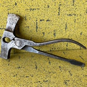 Può includere: Un utensile combinato vintage con testa di martello, pinze e manico curvo. L'utensile in metallo ha un aspetto usurato e invecchiato, con segni di utilizzo visibili. L'utensile è posto su uno sfondo giallo strutturato.