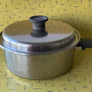 Peut inclure: Une casserole en acier inoxydable avec un couvercle et deux poignées noires. Le couvercle a une poignée noire sur le dessus. La casserole est posée sur une surface jaune.