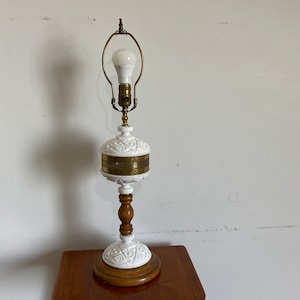 May include: A white milk glass table lamp with a brass shade and a wooden base. The lamp has a white light bulb turned on.
