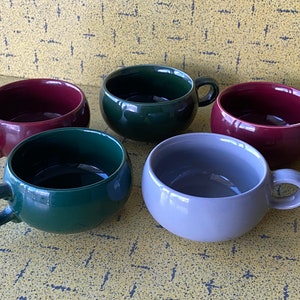 Op de afbeelding: Vijf keramische mokken in verschillende kleuren: bordeauxrood, groen, grijs en nog twee tinten groen. De mokken hebben een afgeronde vorm en een handvat aan de zijkant.