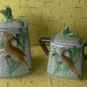 Könnte beinhalten: Ein Set aus zwei Zuckerbehältern aus Keramik mit Deckeln. Die Behälter sind mit einem grünen und braunen Blattdesign und einem stilisierten Hirsch verziert. Die Deckel haben ein grünes Blattdesign.