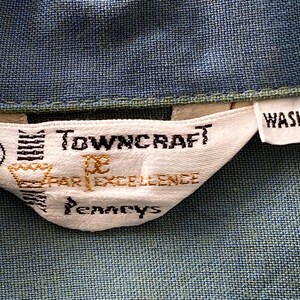 May include: Close-up of a white fabric tag with the text "Towncraft" and "Par Excellence" in black and gold lettering. The tag also includes the word "Washable" and the letter "M". The tag is sewn onto a blue fabric.