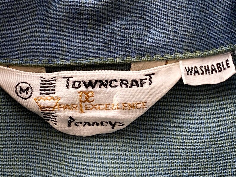 May include: Close-up of a white fabric tag with the text "Towncraft" and "Par Excellence" in black and gold lettering. The tag also includes the word "Washable" and the letter "M". The tag is sewn onto a blue fabric.