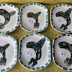 Pode incluir: Seis pratos de cerâmica branca com borda verde e um design de orca estilizado em preto e verde. Cada prato tem um número de 1 a 6 impresso.
