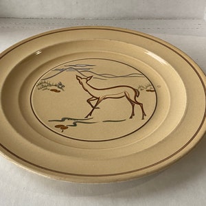 May include: A cream-coloured ceramic plate with a brown rim and a stylised deer design in brown and grey. The deer is standing in a landscape with hills and clouds.