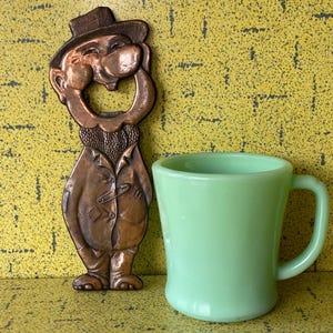 Può includere: Un apribottiglie vintage in rame a forma di uomo sorridente con cappello e cappotto, accanto a una tazza verde chiaro. L'apribottiglie ha un design dettagliato e la tazza ha una forma curva con un manico.