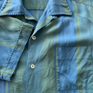 May include: Close-up of a blue and green striped short-sleeved shirt. The shirt has a collar, a front pocket, and white buttons. The fabric appears to be lightweight.