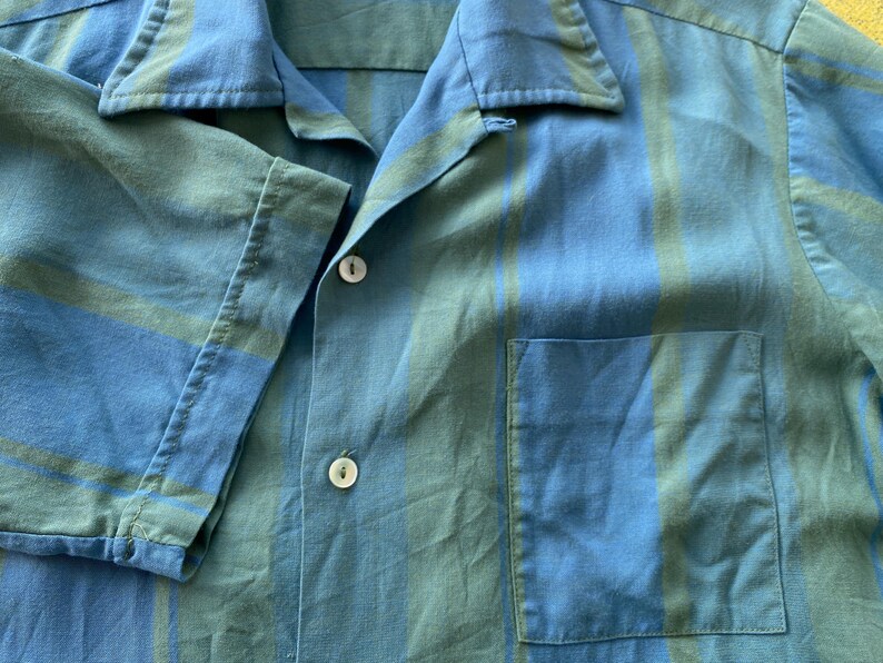 May include: Close-up of a blue and green striped short-sleeved shirt. The shirt has a collar, a front pocket, and white buttons. The fabric appears to be lightweight.