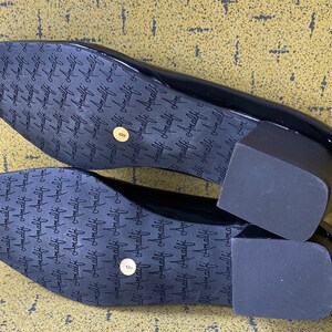 May include: A pair of black patent leather heeled shoes with a square toe. The shoes have a thick block heel and the soles are marked with the word "Amalfi" repeated many times.