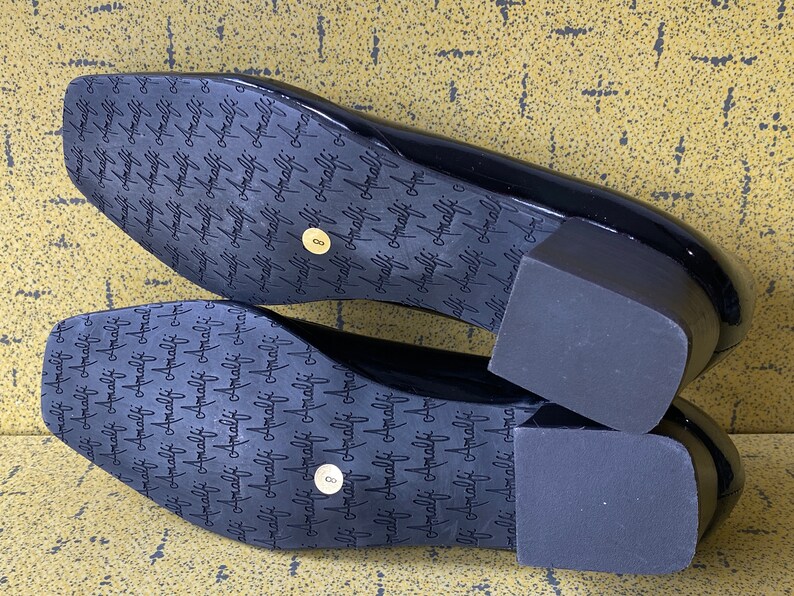 May include: A pair of black patent leather heeled shoes with a square toe. The shoes have a thick block heel and the soles are marked with the word "Amalfi" repeated many times.