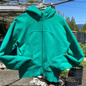 Può includere: Una felpa con cappuccio verde brillante con chiusura a zip. La felpa ha un design curvo distintivo sul davanti.