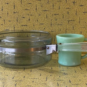 Può includere: Un barattolo di vetro trasparente con una fascia metallica e una tazza verde chiaro con un manico trasparente. Il barattolo è rotondo e ha un bordo. La tazza è piccola e ha un manico. Lo sfondo è giallo strutturato.