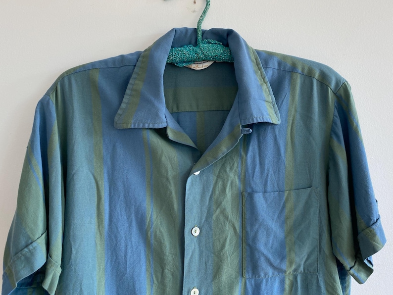 May include: A short-sleeved button-down shirt with vertical stripes in shades of blue and green. The shirt has a collared neckline, a chest pocket, and is hanging on a teal hanger.