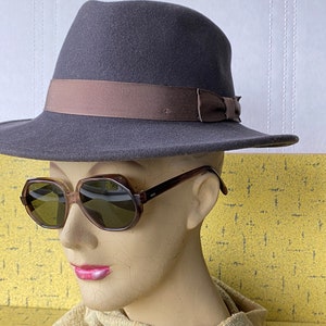 Pode incluir: Um chapéu fedora de feltro cinza com uma fita marrom e um laço. O chapéu é usado em uma cabeça de manequim com óculos de sol de tartaruga marrom.
