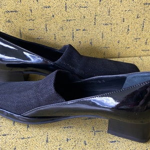 May include: Pair of black heeled shoes with a patent leather upper and a black fabric toe. The shoes have a low block heel.
