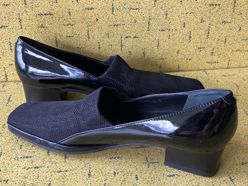 May include: Pair of black heeled shoes with a patent leather upper and a black fabric toe. The shoes have a low block heel.