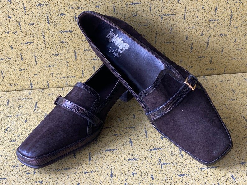 May include: Pair of brown suede heeled loafers with a buckle strap and a square toe. The shoes are labeled "Inglewood by Rampage".
