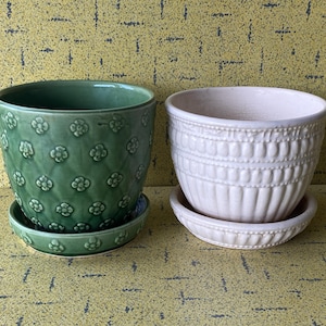 Op de afbeelding: Twee keramische plantenpotten met bijpassende schotels. De linkerpot is saliegroen met een reliëf bloemenpatroon en ruittextuur. De rechterpot is lichtroze met een verticaal geribbeld ontwerp en een kralenrand aan de bovenzijde.