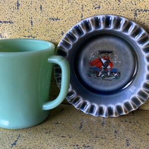 Può includere: Una tazza in ceramica verde con manico si trova accanto a un piattino in ceramica blu e bianco con la raffigurazione di un uomo che porta una grande pentola sulla schiena. Il piattino ha un bordo ondulato e il testo "Fine Macroom" è stampato sul fondo.