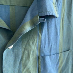 May include: Close-up of a blue and green striped shirt. The shirt has a collar, a pocket, and a white button. The fabric appears to be linen or a similar lightweight material. The color scheme is a mix of light and medium blues with green stripes.