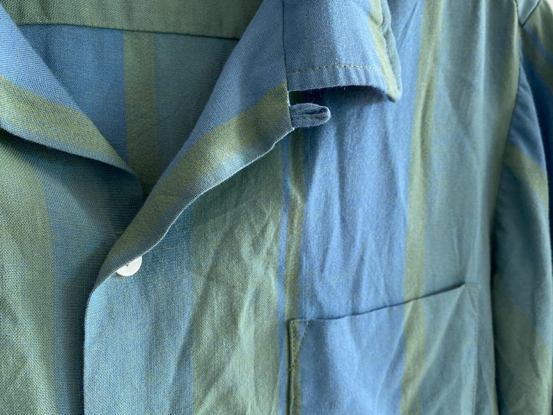 May include: Close-up of a blue and green striped shirt. The shirt has a collar, a pocket, and a white button. The fabric appears to be linen or a similar lightweight material. The color scheme is a mix of light and medium blues with green stripes.