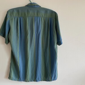 May include: A short-sleeved button-down shirt with a vertical striped pattern in shades of blue and green. The shirt is hanging on a green hanger against a white wall.