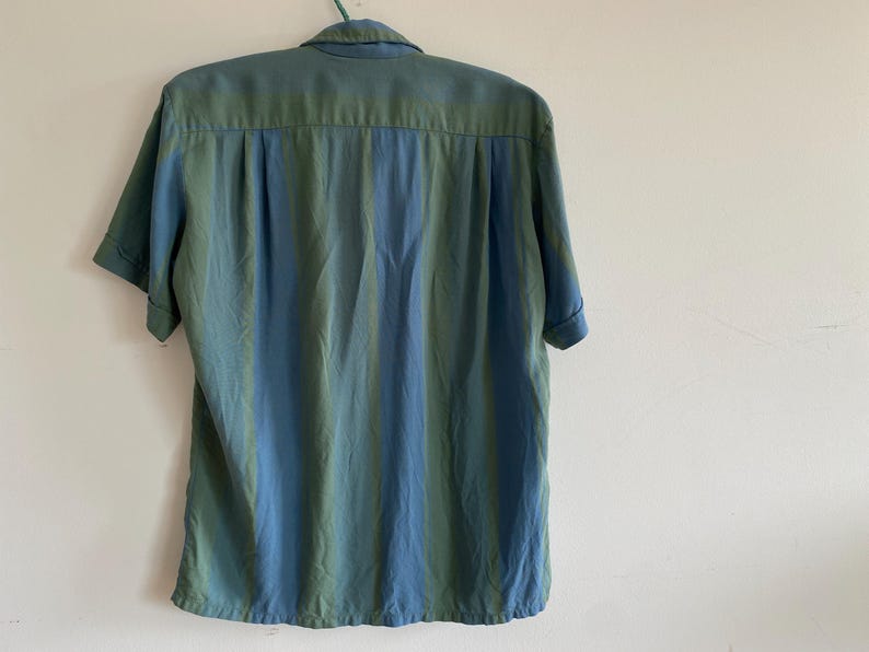 May include: A short-sleeved button-down shirt with a vertical striped pattern in shades of blue and green. The shirt is hanging on a green hanger against a white wall.