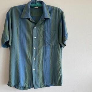 May include: A short-sleeved button-down shirt with vertical stripes in shades of blue and green. The shirt has a classic collar, a chest pocket, and is buttoned up. The garment is hanging on a green hanger against a white wall.