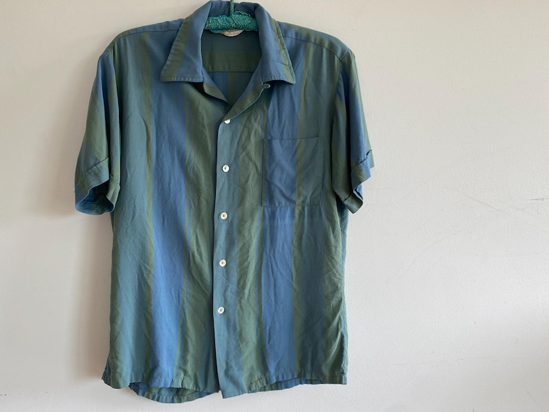 May include: A short-sleeved button-down shirt with vertical stripes in shades of blue and green. The shirt has a classic collar, a chest pocket, and is buttoned up. The garment is hanging on a green hanger against a white wall.