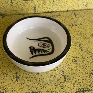 Pode incluir: Uma pequena tigela de cerâmica branca com uma borda preta e um design estilizado de rosto de animal preto e rosa.