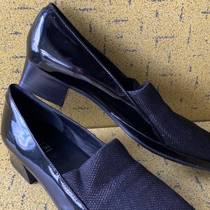 May include: A pair of black patent leather shoes with a low block heel. The shoes have a black fabric upper with a subtle shimmer.