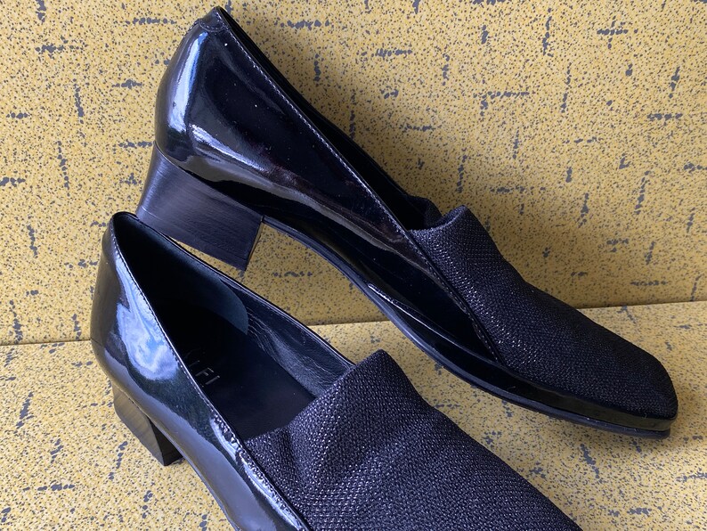 May include: A pair of black patent leather shoes with a low block heel. The shoes have a black fabric upper with a subtle shimmer.